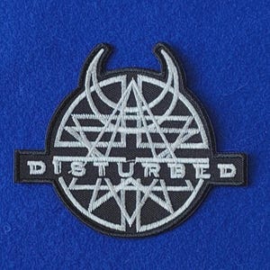 May include: A black and white embroidered patch featuring the word DISTURBED in capital letters. The design includes a circular emblem with geometric lines, triangles, and two curved horn-like shapes at the top, all rendered in white thread.
