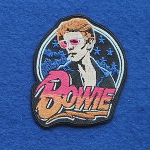 Puede incluir: Un parche bordado que presenta un retrato estilizado de una persona con cabello naranja y gafas de cristales rosados. El diseño incluye un fondo negro con estrellas azules y la palabra Bowie en letras grandes con degradado rosa y naranja.