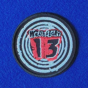 Könnte beinhalten: Ein kreisförmiger gestickter Aufnäher mit schwarzem Hintergrund und konzentrischen hellblauen Ringen. In der Mitte ist der Text Wednesday in Schwarz über einer großen, fetten roten Zahl 13 mit schwarzer Umrandung gestickt, alles schwarz umrandet.