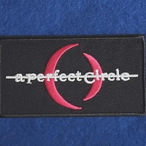 Op de afbeelding: Een rechthoekige zwarte geborduurde patch met een roze halvemaanvormige afbeelding gecentreerd achter de witte kleine letters a perfect circle, met een horizontale witte lijn die door het midden van de tekst en de maangrafiek loopt.