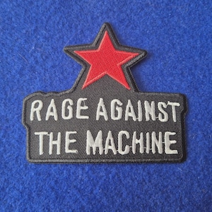Może przedstawiać: Czarna haftowana naszywka z czerwoną gwiazdą u góry oraz białym napisem RAGE AGAINST THE MACHINE w dwóch liniach poniżej. Naszywka posiada czarną obwódkę oraz prostokątną podstawę z kształtem gwiazdy wystającym z górnej części.