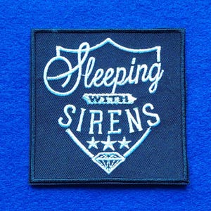 May include: A square black embroidered patch featuring the white text Sleeping with Sirens inside a shield-shaped outline. The design includes three small stars above a diamond graphic at the bottom, all rendered in white thread on a black fabric base.