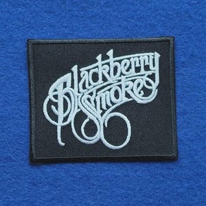 May include: A rectangular black embroidered patch featuring the white text Blackberry Smoke in a stylised, cursive font. The patch has a black stitched border and is designed for application onto fabric items like jackets, bags, or denim clothing.