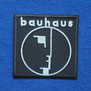 May include: A square black embroidered patch featuring the white text "bauhaus" at the top and a white circular line drawing of a stylised human face profile centred below the text, with a black stitched border surrounding the entire design.