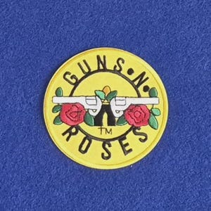 May include: A circular yellow patch featuring the Guns N' Roses logo. The design includes two white revolvers crossed behind a central green leaf, flanked by two red roses with green leaves. The text GUNS N' ROSES is printed in black.