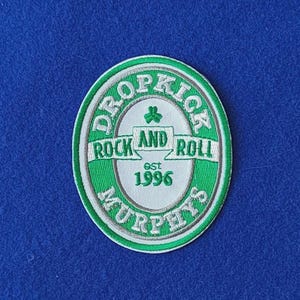 Może przedstawiać: Owalna haftowana naszywka z zielono-białym wzorem. Tekst brzmi DROPKICK MURPHYS z centralnym banerem z napisem ROCK AND ROLL oraz tekstem est 1996 pod małą ikoną zielonej koniczyny. Obramowanie jest biało-zielone.