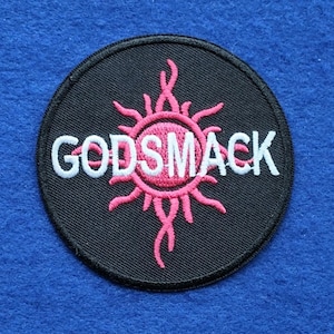 May include: A circular embroidered patch featuring a black background with a pink sun-like graphic at the center. The word GODSMACK is stitched in white capital letters across the middle of the design, which is finished with a black stitched border.