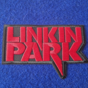 May include: A rectangular embroidered patch featuring the text LINKIN PARK in bold red lettering. The letters are stitched onto a black background with a black border, and the design includes a sharp, angular extension pointing downward from the letter K.