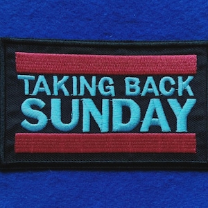 Puede incluir: Un parche rectangular bordado con fondo negro que presenta el texto TAKING BACK SUNDAY en letras mayúsculas de color azul claro. El diseño incluye dos franjas horizontales rojas situadas encima y debajo de las letras azules.