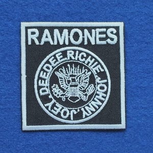 Peut inclure: Un écusson brodé carré noir et blanc avec le texte RAMONES en haut. En dessous, un emblème circulaire affiche un graphique d'aigle entouré des noms DEEDEE, RICHIE, JOHNNY et JOEY dans une police de caractères blanche brodée.