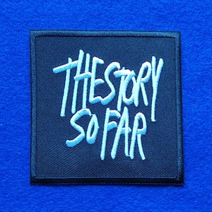 May include: A square black embroidered patch featuring the text THE STORY SO FAR in white thread. The lettering is stylised in a handwritten font, and the patch has a clean, stitched border, creating a distinct contrast against the dark fabric.