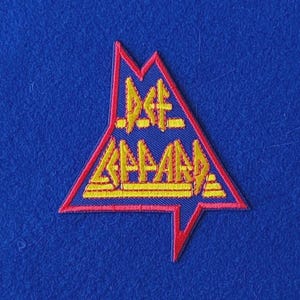 May include: A triangular embroidered patch featuring the text Def Leppard in yellow thread. The patch has a blue interior background and a red border. The design is shaped like a stylised arrow pointing upwards, with the band name clearly visible.