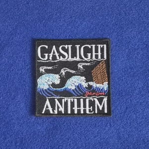Op de afbeelding: Een vierkante zwarte geborduurde patch met de witte tekst GASLIGHT ANTHEM. Het ontwerp toont gestileerde blauwe en witte oceaangolven, een bruine houten structuur en kleine witte figuren tegen een zwarte achtergrond met een gestikte rand.