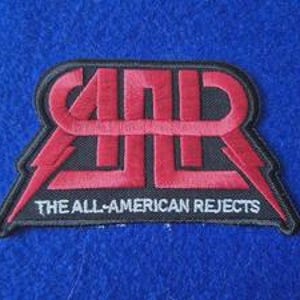 May include: A black embroidered patch featuring the red stylised AAR logo with lightning bolt accents on the sides. Below the logo, the text THE ALL-AMERICAN REJECTS is stitched in white thread. The patch has a black border and a textured finish.