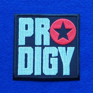 May include: A square embroidered patch with a black background and light blue block lettering that spells out PRODIGY. The letter O is replaced by a red circular emblem containing a black five-pointed star. The patch has a black stitched border.