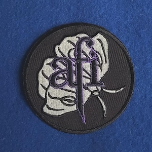 May include: A circular embroidered patch featuring a black background with a white stylized butterfly graphic. The letters afi are embroidered in purple over the center of the butterfly design, with a black border surrounding the entire circular shape.