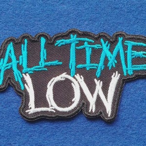 May include: A black embroidered patch featuring the text ALL TIME LOW. The words ALL TIME are stitched in bright cyan thread with a distressed, jagged font style, while the word LOW is stitched in white thread with a similar jagged aesthetic.