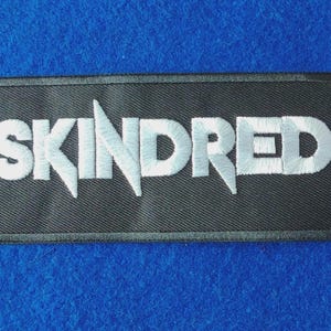 May include: A rectangular black embroidered patch featuring the word SKINDRED in bold white capital letters. The lettering has a distinct stylised font with sharp, pointed edges on the letters S, K, I, N, and D, all set against a black fabric background.