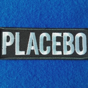 May include: A rectangular embroidered patch featuring a black background with the word PLACEBO stitched in bold, white, capital letters. The patch has a clean, stitched border and a flat, fabric texture throughout the design.