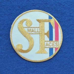 Könnte beinhalten: Ein kreisförmiger bestickter Aufnäher mit den gold gestickten Buchstaben S und F auf weißem Hintergrund. Das Wort SMALL ist in Gold zwischen die Buchstaben gestickt, das Wort FACES in Gold darunter. Ein blauer und roter Streifen säumt die rechte Seite.