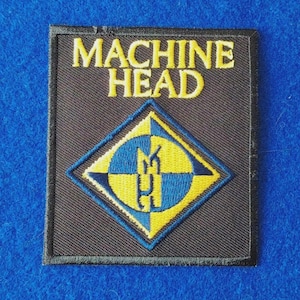 May include: A rectangular black fabric patch featuring the yellow embroidered text MACHINE HEAD at the top. Below the text is a diamond-shaped emblem with a blue and yellow checkered pattern and the stylised letters MH in the centre.