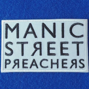 Op de afbeelding: Een rechthoekige geborduurde patch met een witte achtergrond en zwarte tekst. De tekst luidt MANIC STREET PREACHERS, waarbij de letters S in STREET en de letters P, R, E, A, C, H, E, R, S in PREACHERS in spiegelbeeld zijn weergegeven.