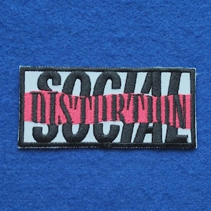 Peut inclure: Un écusson rectangulaire brodé présentant le texte SOCIAL DISTORTION en lettres noires. Le mot SOCIAL est brodé en noir sur un fond gris clair, tandis que le mot DISTORTION est brodé en noir sur une bande horizontale de couleur rose.