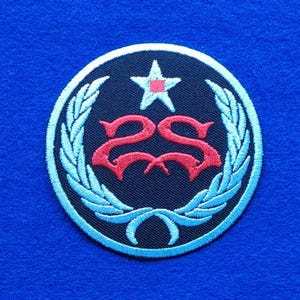 Op de afbeelding: Een ronde geborduurde patch met een donkerblauwe achtergrond en een lichtblauwe lauwerkransrand. In het midden bevindt zich een rood gestileerd dubbel S-monogram onder een lichtblauwe ster met een klein rood vierkant in het midden.