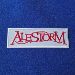 May include: A rectangular embroidered patch featuring the word ALESTORM in red thread with a stylised font. The lettering is set against a light grey background with a white border, creating a clear contrast for the band logo design.