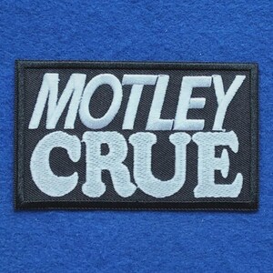 May include: A rectangular embroidered patch featuring the text MOTLEY CRUE in bold white capital letters against a solid black background. The patch has a thick black stitched border and a textured fabric finish throughout the design.