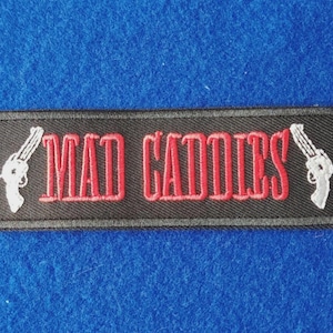 Peut inclure: Un écusson rectangulaire brodé noir avec le texte rouge MAD CADDIES en police grasse, flanqué d'un revolver blanc brodé sur les côtés gauche et droit, le tout entouré d'une bordure noire cousue sur tout le contour de l'écusson.