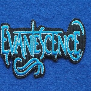 May include: An embroidered fabric patch featuring the word EVANESCENCE in a stylised, jagged font. The lettering is stitched in bright blue thread with a black background, creating a high-contrast design with a distinct, sharp, and angular aesthetic.