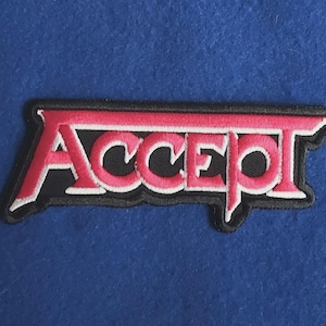 May include: A rectangular embroidered patch featuring the word ACCEPT in bold, stylized pink lettering with a white outline, set against a solid black background. The text has a distinct, sharp font style with a horizontal bar across the top.