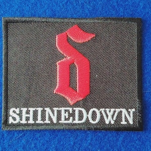 May include: A rectangular black embroidered patch featuring a red stylised letter S logo centred above the white text SHINEDOWN. The patch has a black stitched border and a textured fabric surface throughout the design.