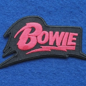 May include: An embroidered patch featuring the word Bowie in bright pink thread against a black background. The design includes a stylized lightning bolt shape integrated into the lettering, with a jagged, asymmetrical border surrounding the text.