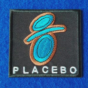 May include: A square embroidered patch with a black background featuring a stylised blue and brown logo design. The word PLACEBO is stitched in white capital letters at the bottom of the patch. The entire design is bordered by a black stitched edge.