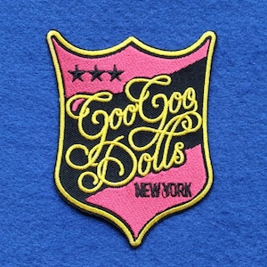 May include: A shield-shaped embroidered patch featuring a pink and black background with a yellow border. The text Goo Goo Dolls is stitched in yellow cursive script, with three black stars above and the word NEW YORK in black below.