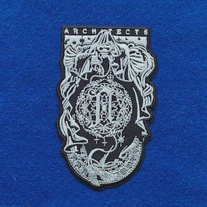 Op de afbeelding: Een zwart-witte geborduurde patch met het woord ARCHITECTS bovenaan. Het ontwerp toont een skeletachtige figuur met een kap boven een rond embleem met een sierlijke letter N in het midden, omringd door ingewikkelde filigraan en kruismotieven.