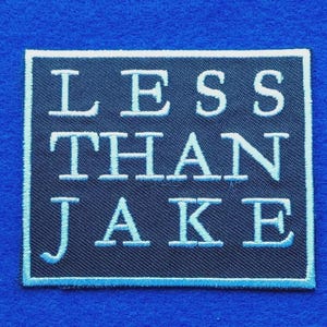 Pode incluir: Um emblema bordado retangular com um fundo azul-marinho escuro e uma borda azul-clara. O texto LESS THAN JAKE está bordado em letras maiúsculas azul-claras no centro do emblema com uma fonte de estilo serifado.