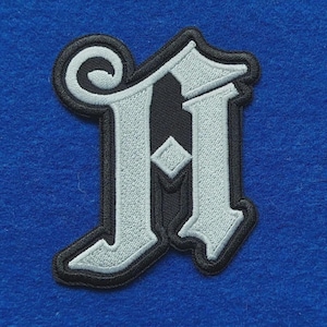 May include: An embroidered patch featuring the letter A in a gothic font style. The letter is light grey with a black border and a black inner section, featuring a decorative swirl at the top left and a small diamond shape in the centre.