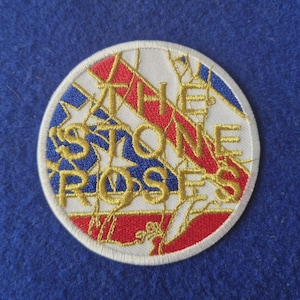 May include: A circular embroidered patch featuring the text THE STONE ROSES in gold thread. The background design consists of a white field with red and blue diagonal stripes and a gold-stitched abstract pattern resembling cracked paint.