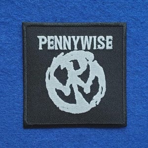 May include: A square black fabric patch featuring the white embroidered text PENNYWISE at the top and a circular white embroidered logo design in the centre. The patch has a black stitched border and a flat, woven texture throughout the design.