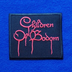 Op de afbeelding: Een rechthoekige zwarte stoffen patch met de tekst Children Of Bodom geborduurd in een gestileerd roze lettertype. De letters hebben een druipend effect, met dunne lijnen die naar beneden lopen vanaf de tekens op een donkere achtergrond.