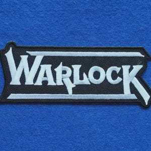 May include: A rectangular embroidered patch with a black background and a white border. The word WARLOCK is stitched in white capital letters across the centre, framed by horizontal white lines that extend to the edges of the patch shape.