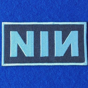 May include: A rectangular embroidered patch featuring a black background with the letters NIN stitched in light blue. The letters are arranged horizontally, with the final N appearing in a mirrored orientation. The patch has a light blue border.