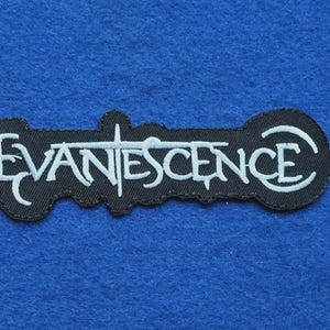 May include: An embroidered patch featuring the word EVANESCENCE in white thread on a black background. The lettering uses a stylised font with a cross integrated into the letter T, and the entire design is outlined in a black border.