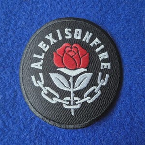 Peut inclure: Un écusson brodé noir de forme circulaire représentant une rose rouge avec des feuilles blanches et une chaîne blanche enroulée autour de la tige. Le texte ALEXISONFIRE est brodé en lettres capitales blanches sur le bord supérieur.