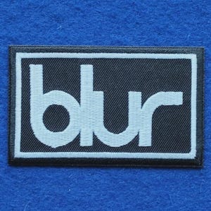 May include: A rectangular embroidered patch featuring a black background with a white border and the word blur written in white lowercase letters. The patch has a stitched texture and is designed for application onto fabric surfaces.
