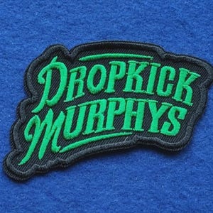 May include: A black embroidered patch featuring the text DROPKICK MURPHYS in bright green thread. The lettering is stylised in a bold, cursive font with a thick black border outlining the shape of the words, creating a distinct contrast.