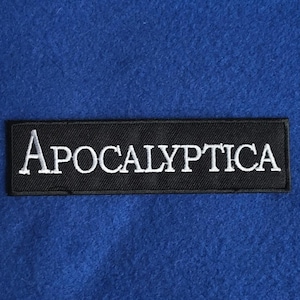 以下が含まれることがあります： スタイリッシュなフォントで白い大文字の「Apocalyptica」という単語が刺繍された、長方形の黒いワッペン。ワッペンには黒い縁取りがあり、柔らかく毛羽立った質感の鮮やかな青い布地を背景に配置されています。